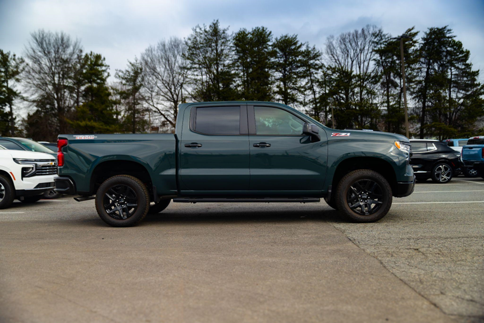 Side view of a Chevy Silverado four-door pickup truck parked in a dealership lot, with trees and other vehicles in the background.
