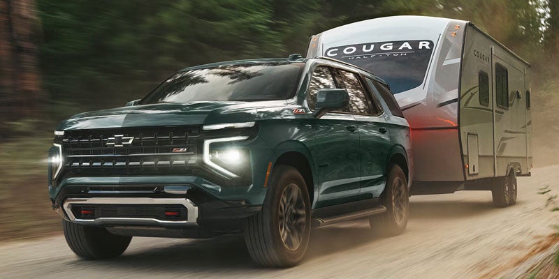 A dark green Tahoe towing a Cougar travel trailer along a dusty road through a forest.